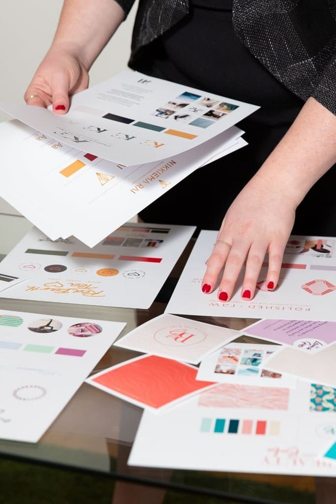 egy pirosra festett körmű női kéz színek és minták között válogat, melyek szét vannak terítve az íróasztalon. másik kezében egy újabb kupac papír tele mintákkal
